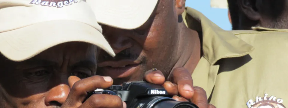 Two dark skinned men stand close together, staring at a digital camera screen. They are wearing tan baseball caps and tan buttons up shirts that say "Rhino Rangers" on the lapel. The back of another man's head is visible in the background.