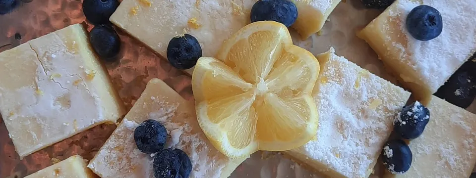 A display of lemon bars with a carved lemon wedge on top and blueberries sprinkled around them.