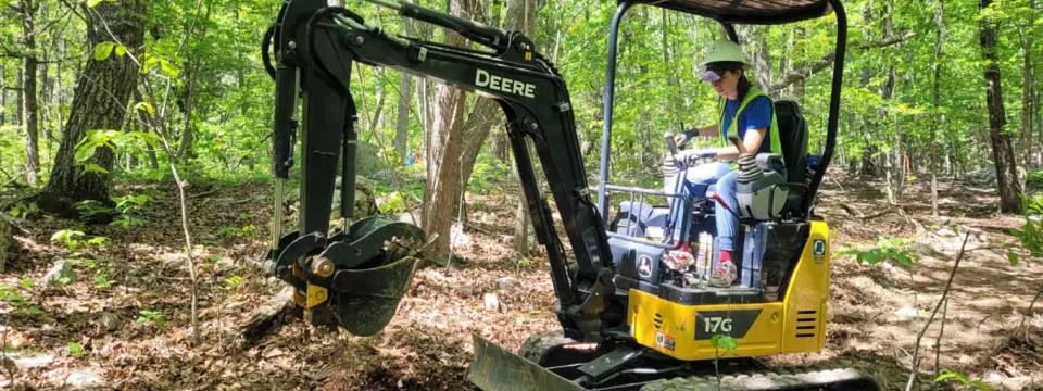 A person wearing protective gear driving a small, yellow construction vehicle with a digging bucket on the front in the woods. They appear to be scooping up dirt to clear the space.