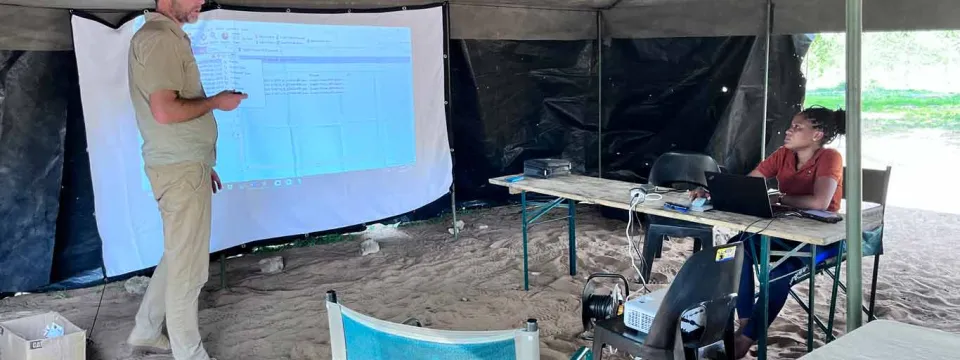 A man in a tan uniform stands to the left, gesturing towards a white projection screen displaying text and data. The screen is set up inside a large, open-sided tent with a sandy floor. To the right, a woman sits at a wooden table with a laptop, looking at the man. A projector is on a chair in front of the table. Camping chairs and another table with a blue notebook are also visible in the foreground.