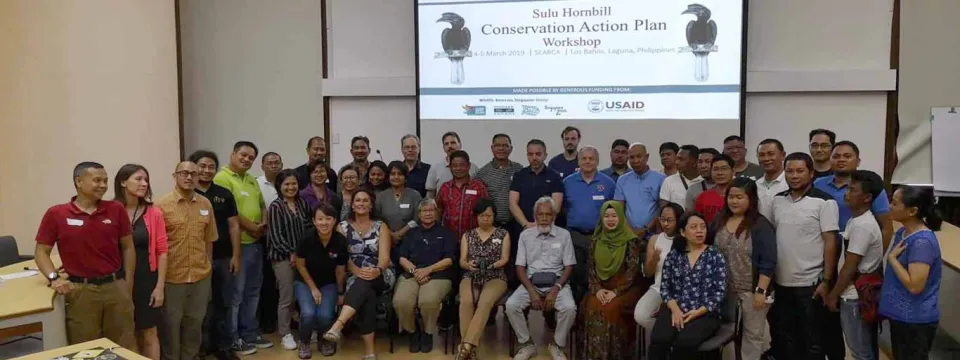 A photo of a large group of diverse participants at the "Sulu Hornbill Conservation Action Plan Workshop," standing and sitting for a group picture in front of the presentation screen.