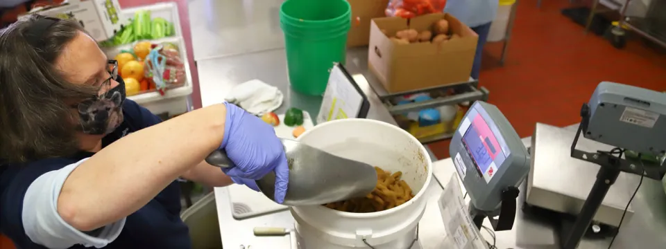A woman, wearing a face mask and blue latex gloves using a metal scoop to transfer food from a work surface into a large white bucket. She is preparing animal feed in a clean, industrial-style kitchen or prep area. Various fruits and vegetables are visible in the background, and to the right, a digital scale is used for weighing ingredients.
