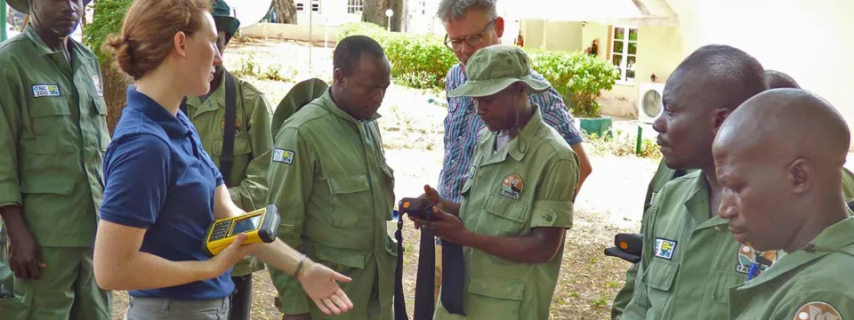 A white woman with a brown ponytail, wearing a dark blue polo shirt and khaki pants, stands on the left, holding a yellow handheld device and gesturing toward a group of men. The majority of the men are wearing uniform green park ranger uniforms, and are looking intently at an item one of them is holding. A white man with gray hair and glasses is standing in the background, observing.