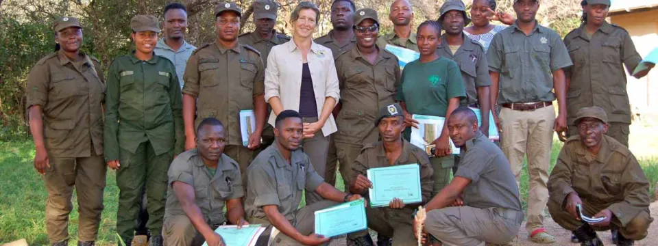 A group of people, mostly uniformed park rangers, poses for a photo outside. A woman in a white blazer and black pants stands in the center of the back row, and two men kneeling in the front row are holding up certificates.