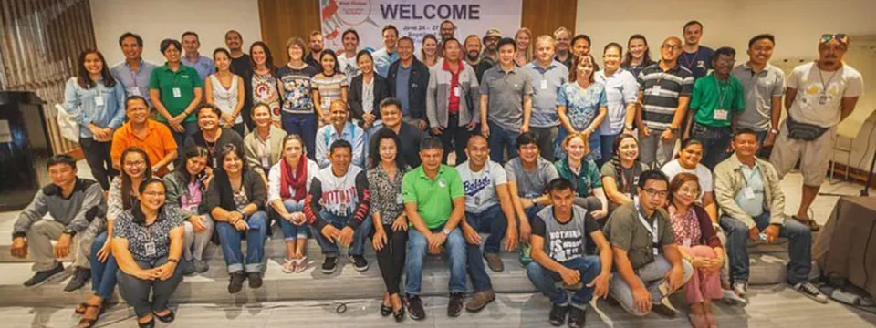 A large group of approximately 30-40 people, likely participants in a workshop or conference, are gathered for a group photo, with some standing in the back and others seated in the front row. A banner is visible on the wall behind them.
