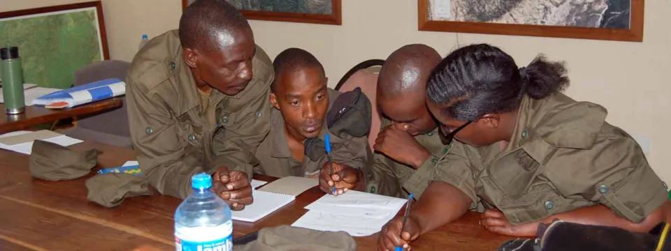 Four people wearing park ranger-style uniforms are gathered around a wooden table in an office. They are all looking down and appear to be discussing documents on the table. The person on the far right, a woman with her hair pulled back, is writing with a blue pen. Two men on the left and a third man next to the woman are leaning in to observe the documents. A bottle of water sits on the table, and two framed maps hang on the tan wall in the background.