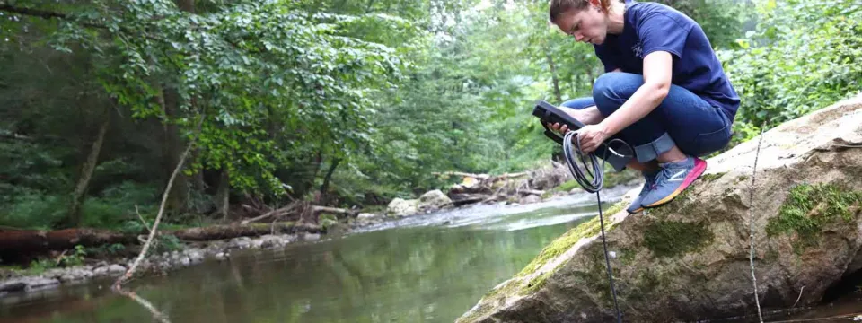 A female field researcher is kneeling on a large rock at the edge of a slow-moving, wooded stream or river. She is wearing a dark t-shirt and jeans and is using a handheld electronic multi-meter to measure water quality. A black cable runs from the device into the dark water. The banks of the stream are densely covered with green trees and foliage.