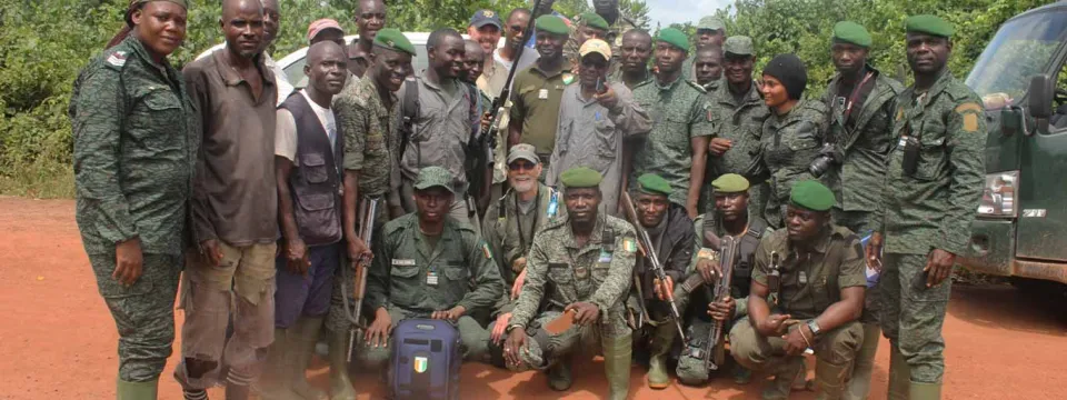 A group of approximately fifteen individuals, most wearing military or camouflage uniforms, pose for a group photo outdoors. They are standing and kneeling on a dirt road, with trees and a dark vehicle visible in the background.