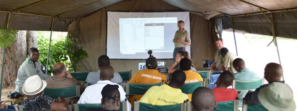 A large outdoor canvas tent, where a group of people are attending a presentation or training session. A man in a khaki shirt is standing under a projected screen on the back wall, speaking to the seated audience. About a dozen individuals, mostly men, are seated on metal-framed chairs, facing the front.