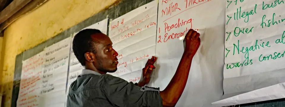 A man in a gray shirt stands, writing on a chalk board that has been covered with large white sheets of poster paper.