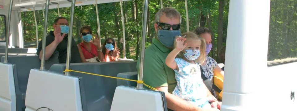 Several families with young children sit spaced apart and wearing face masks on an outdoor tram, waving at the viewer.