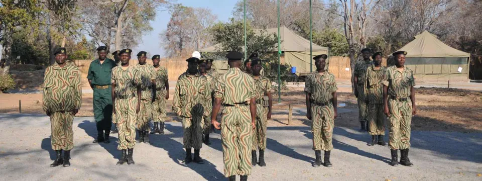 A group of about seven people dressed in uniform (tan shirts and pants with dark boots and hats), possibly park rangers. They are standing in a straight formation on a paved area. One person, on the left, appears to be wearing a darker green uniform. In the background, there are two large light-colored canvas tents and scattered trees with sparse foliage, suggesting a dry or temporary encampment setting.