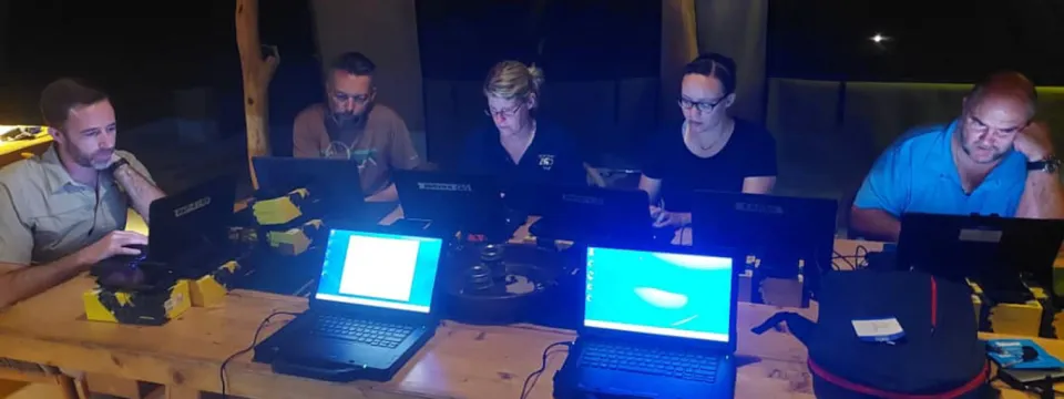 Three men and two women—are seated shoulder-to-shoulder at a long wooden table in a dimly lit, outdoor tent setting, working intently on their individual laptops. They appear to be a research or field team, concentrating on their screens late at night, with various equipment bags on the table.