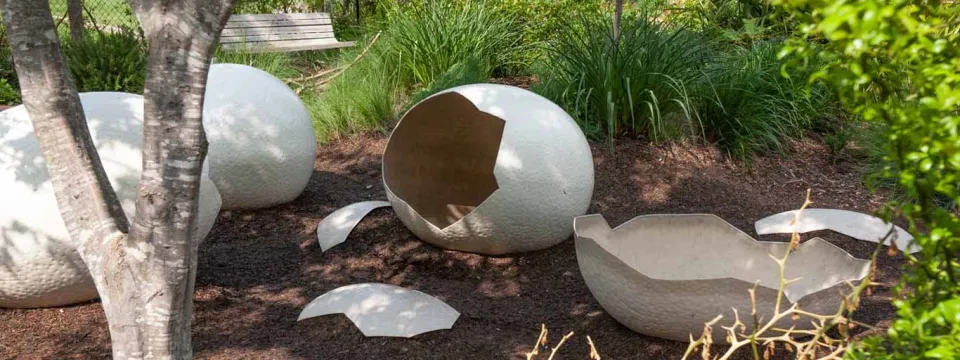 Three oversized, cracked white eggshells are scattered on a bed of dark mulch or dirt in a garden setting, suggesting a recent "hatching." The shells have a textured surface like rough concrete or stone. The scene is surrounded by lush green foliage and tall grass, with the base of a small tree in the foreground to the left and a wooden bench visible in the background. This is likely an outdoor art installation or a zoo/garden exhibit.