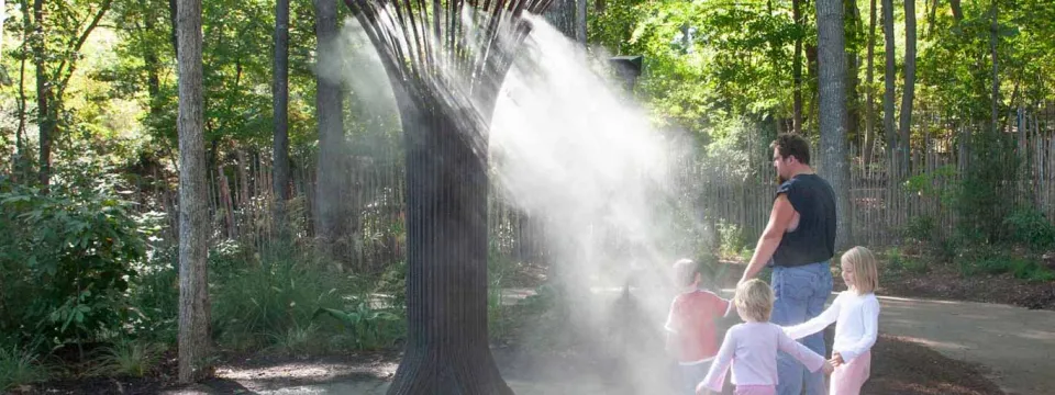 An outdoor misting station, which is a tall, sculpted metal structure resembling a tree, spraying a fine mist to cool visitors. A man and three children are standing near the structure on the right, enjoying the mist. The ground is paved, and the area is surrounded by a dense forest of tall trees and undergrowth.