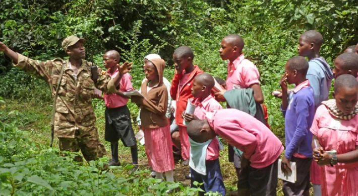 A group of children, most appearing to be Black, are gathered in a grassy area with dense foliage, listening to a person in a military-style uniform who is gesturing and pointing. The children are wearing various colored tops and dresses. The children are students from the UNITE program on a field trip.