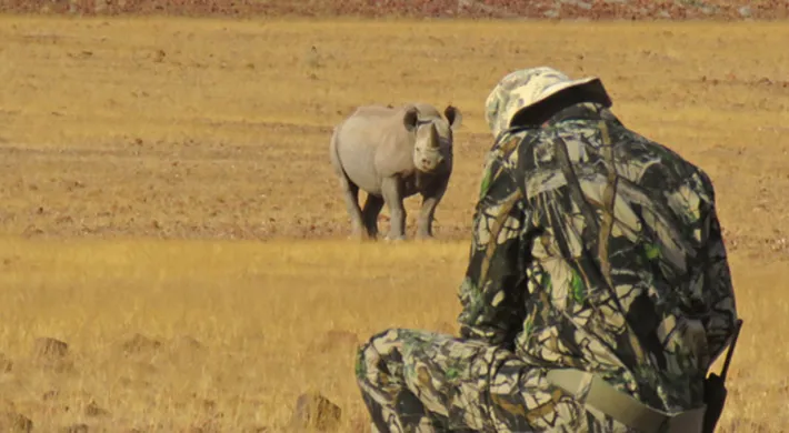 A person wearing camouflaged colored clothes crouched down conducting a rhino monitoring with Save the Rhino Trust. There is a rhinoceros in the background facing the person in a savanna environment.