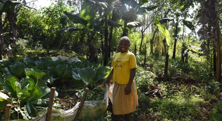 A person with dark skin and a shaved head stands smiling in a sunny, lush green garden. They are wearing a bright yellow t-shirt and a tan skirt. To their left, large, leafy green plants are growing in rows in some light-colored sacks that have been repurposed for gardening. Tall trees and various green plants fill the background, suggesting a tropical environment.