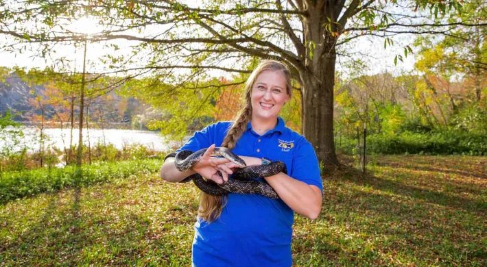 A woman with a long, blonde braid, wearing a blue collared shirt is standing next to a lake in the woods. She is smiling at the camera and holding a very long, black snake in both hands as it wraps around her hands and forearms.