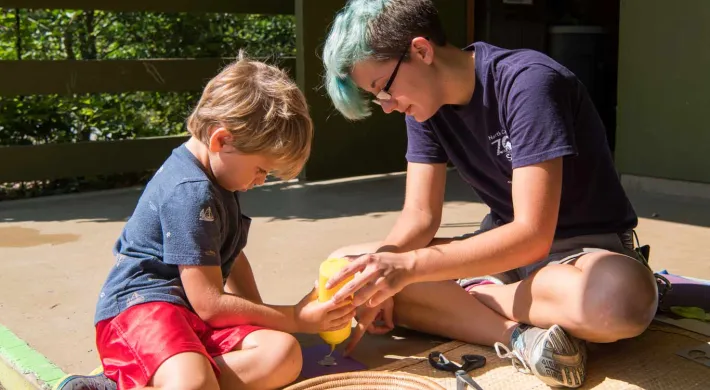 An adult with short blue hair sits on the ground next to a child. They are holding a bottle of what appears to be glue and seem to be working on a craft together on a covered porch area.