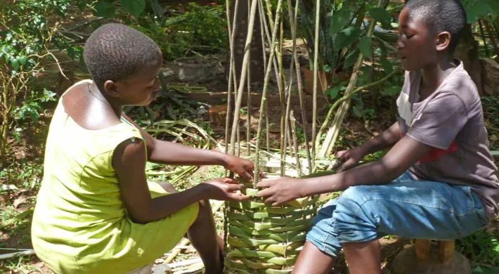 Two children are sitting on the ground outdoors, engaged in weaving a tall basket from natural, green plant material. They are working together to interlace the horizontal strands around the vertical wooden stakes that form the frame of the basket. The scene is set in a sunny, forested area.