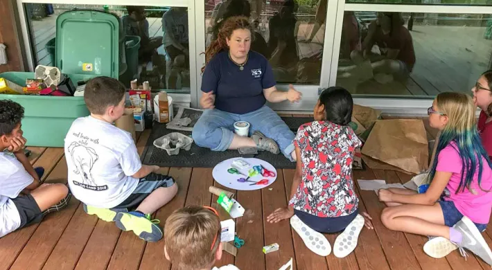 A teacher is leading a group of six children sitting on a wooden porch, demonstrating a craft project using repurposed materials like cardboard and egg cartons.