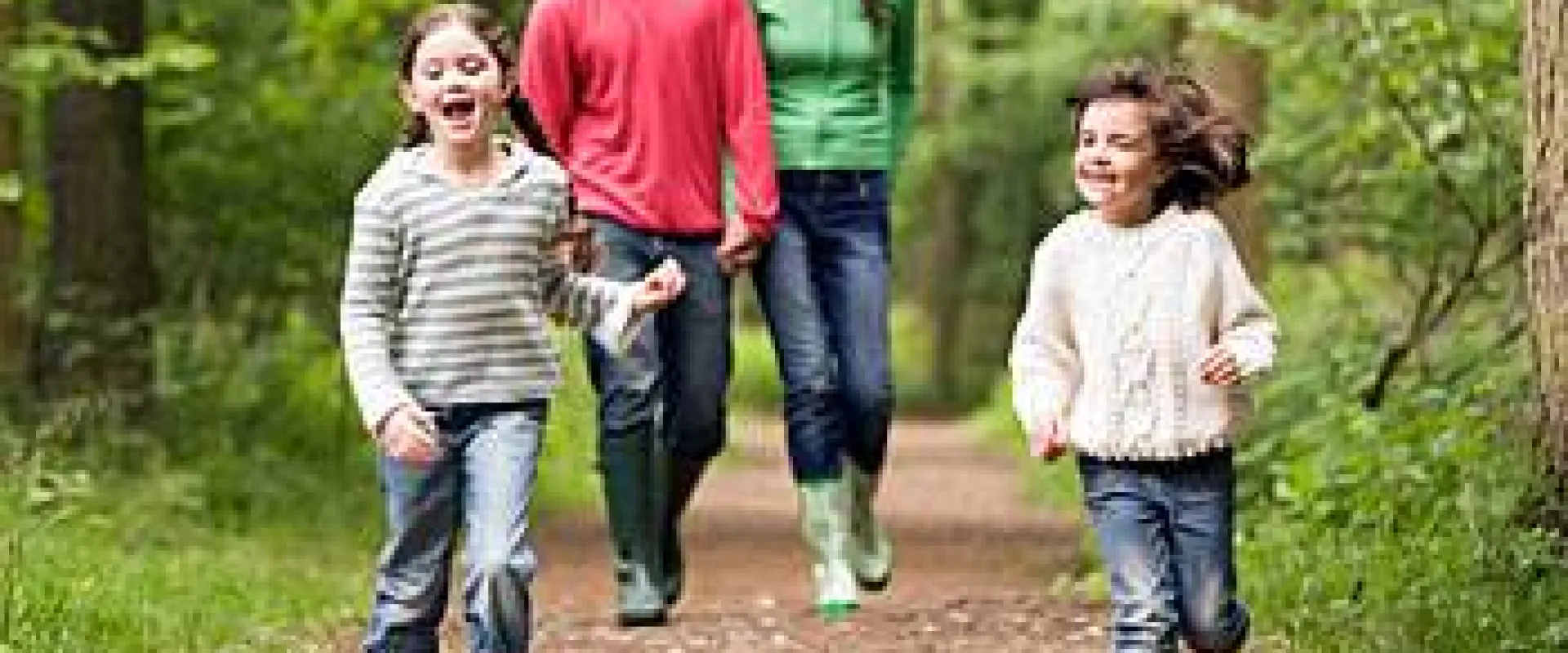 A group of four people, possibly a family, consisting of a man, a woman, and two small girls, walks along a dirt path in a wooded area. The girls are running ahead of the adults and appear to be laughing. The adults are walking behind them and may be smiling. The individuals are wearing casual clothing, including jeans and sweaters.