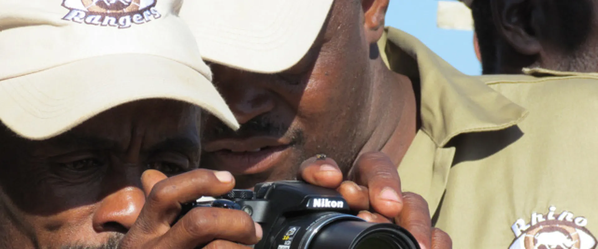 Two dark skinned men stand close together, staring at a digital camera screen. They are wearing tan baseball caps and tan buttons up shirts that say "Rhino Rangers" on the lapel. The back of another man's head is visible in the background.