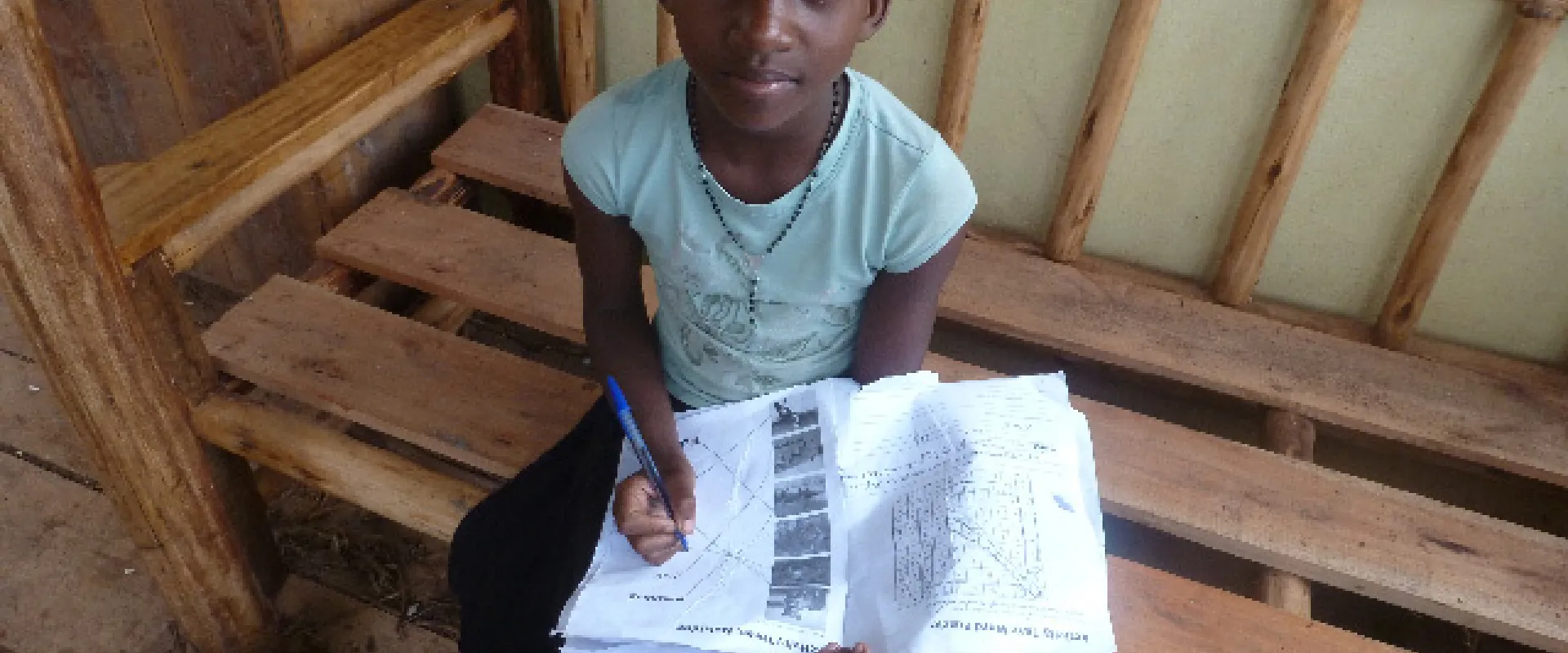 A young child sits on a wooden bench, holding a notebook and pen and looking up at the camera. They have dark skin, short hair, and are wearing a light blue shirt and a black skirt.