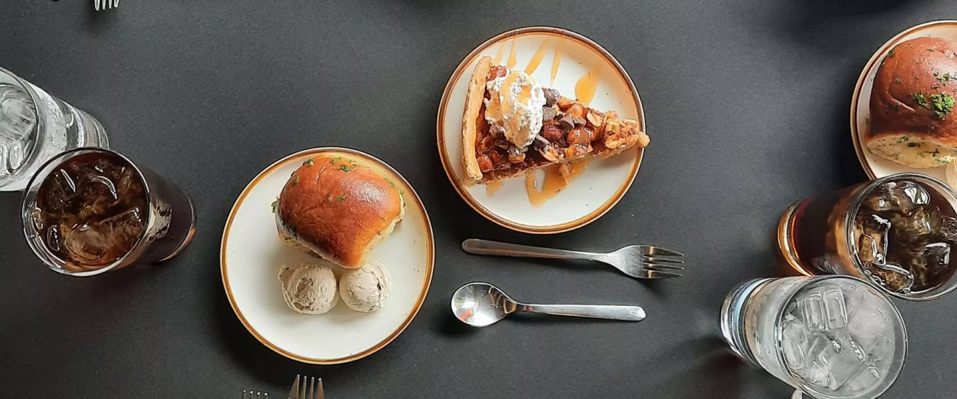 A spread of various foods on plates including as pie, sandwiches, and drinks on a grey table with cutlery sprinkled throughout.