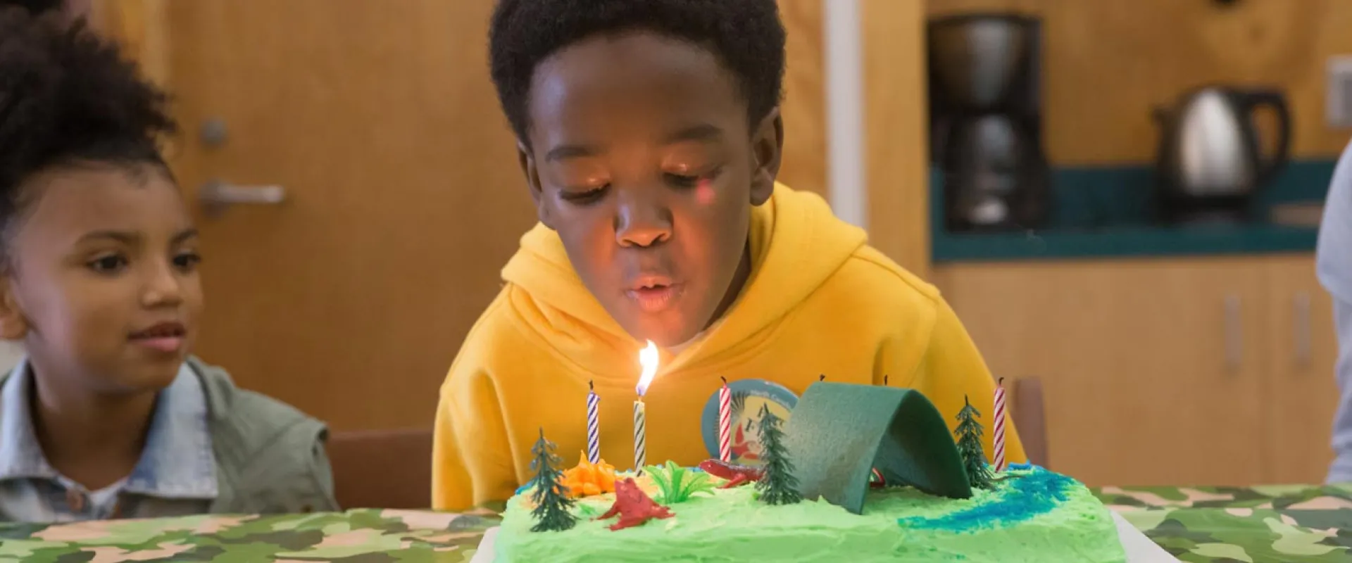 A young child sits at a decorated table, blowing out a candle on a birthday cake. There is another child sitting next to him and a door, sink, and counter with a kettle and a coffee pot on it in the background.