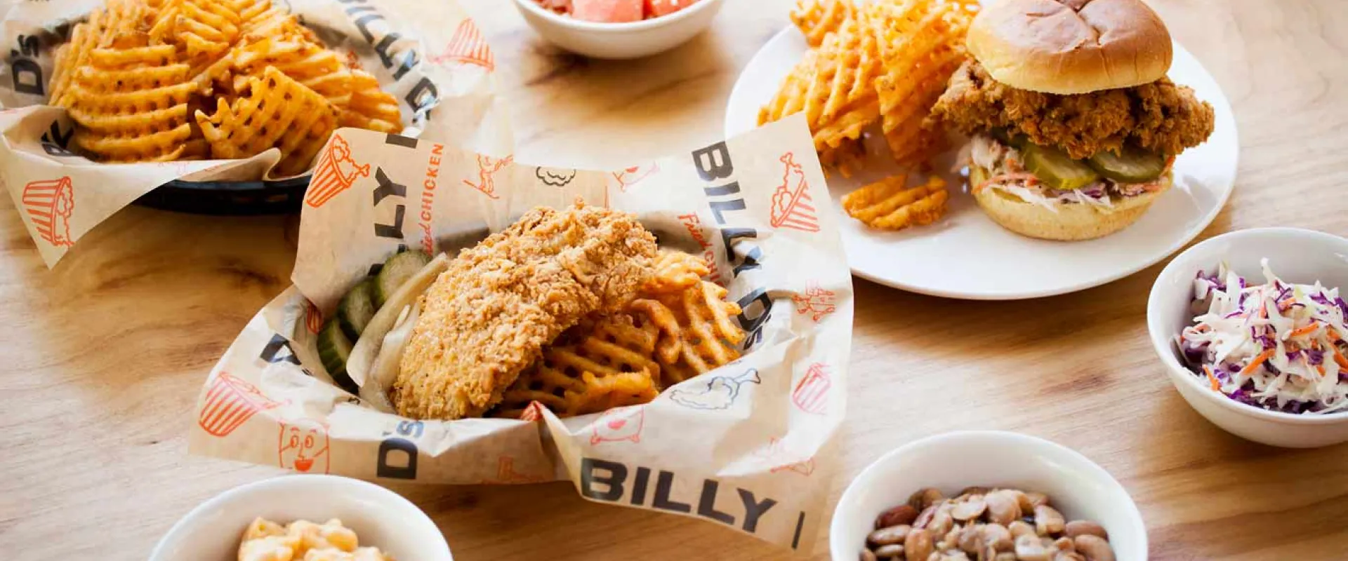 A wooden table with several plates heaped full of food including french fries, fried chicken, and cole slaw.