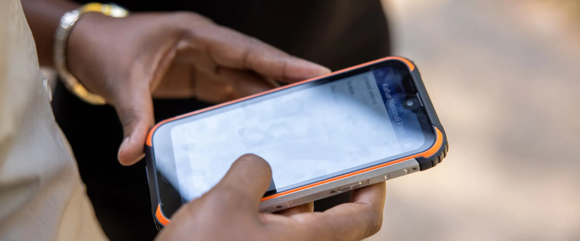 A pair of hands holding a device that looks like a cellphone. Another person, visible from the waist down, wearing black pants, is standing next to them.