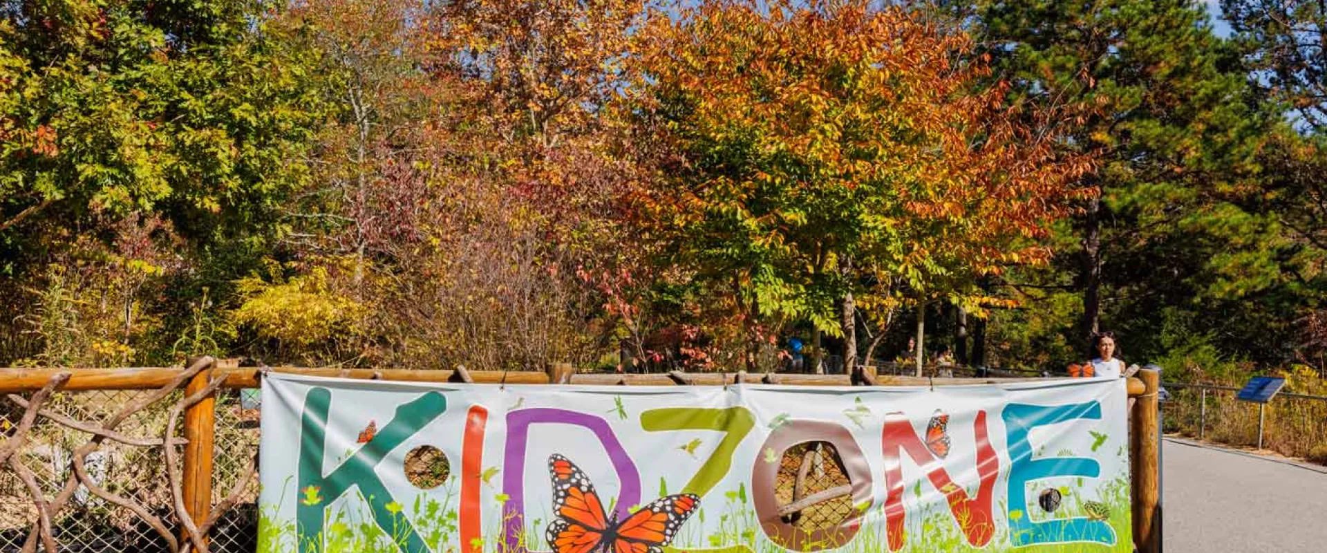 A natural wooden fence covered in vines in front of a wooded area. Attached to the fence is a colorful banner with the text "Kidzone" and images of grass and butterflies.