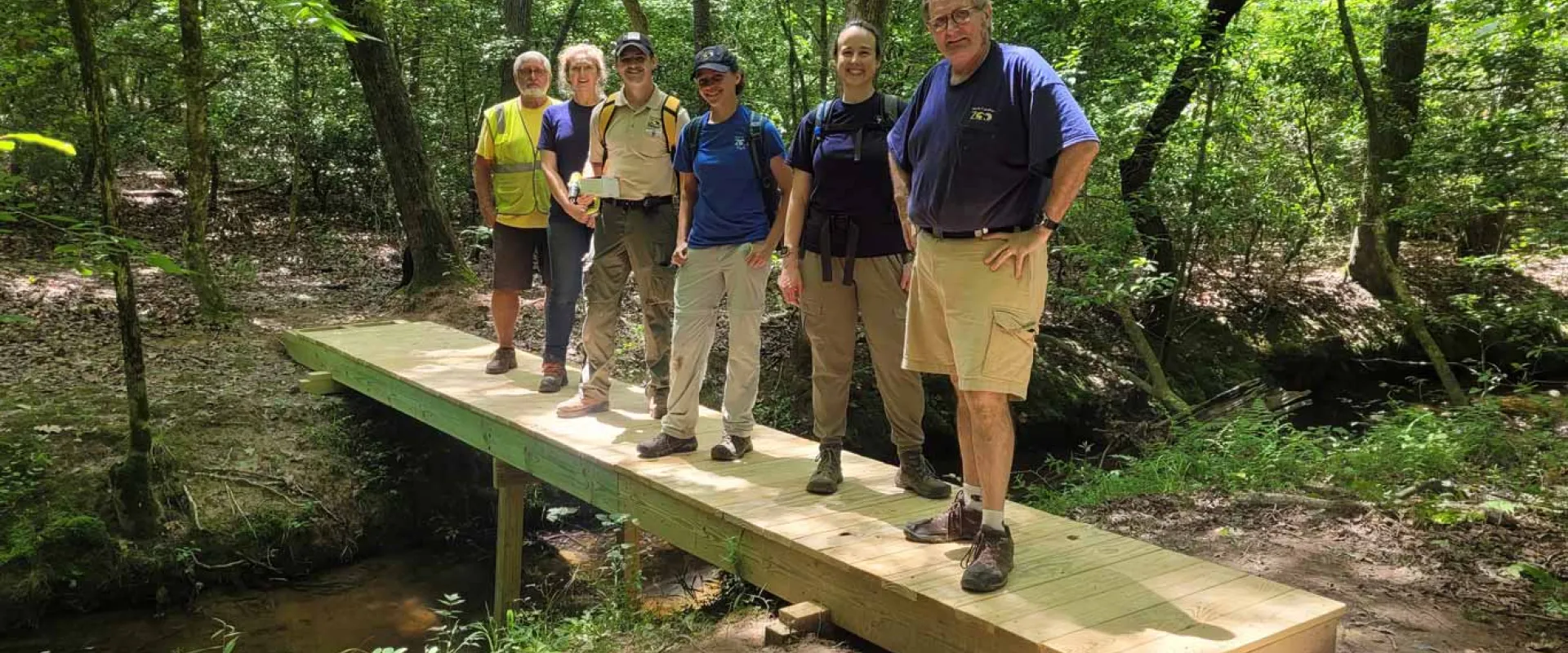 Trail building team consisting of six people of varying ages, races and genders, standing on a bridge that spans a small creek in the woods. They are standing in a line and posing for a photo.