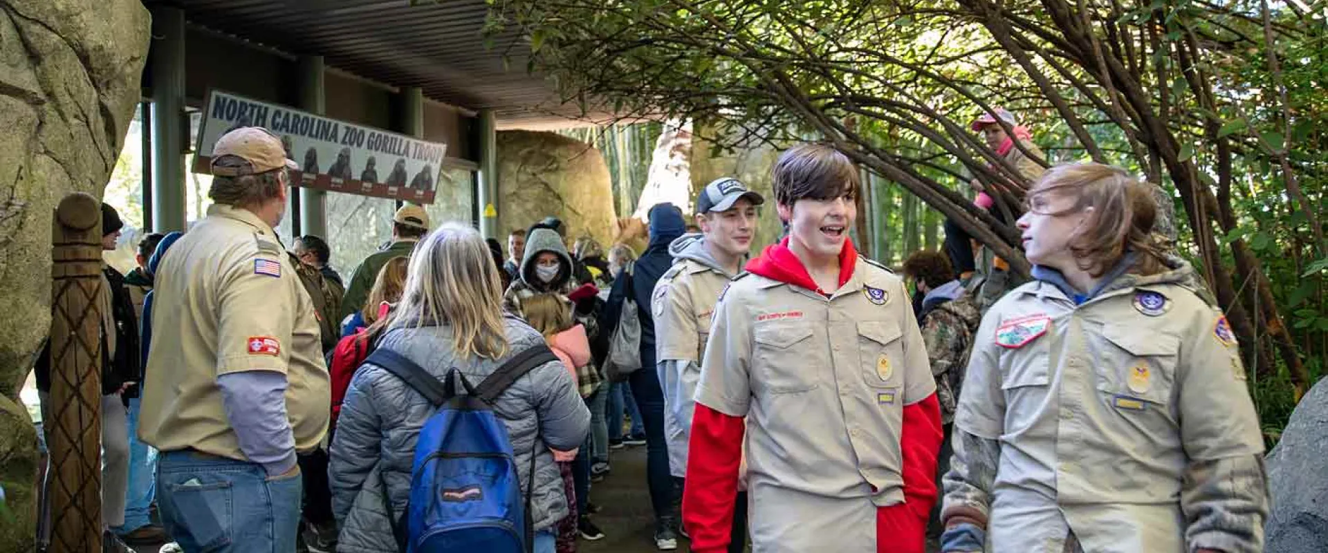 A large group of people with varying ages gathered in front of and walking by a large window and rock wall. They are all wearing Boy Scout uniforms consisting of jeans and tan collared shirts with patches on them.