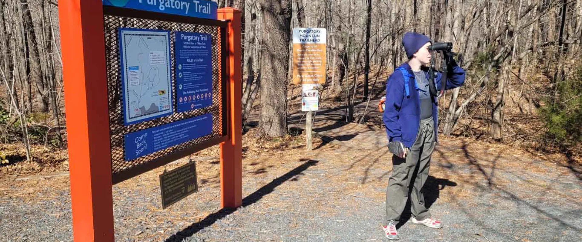 A person in cold weather gear, stands on a trail in the woods, looking off to the right with a pair of binoculars. Next to them is a large sign with the title "Purgatory Trail" with a map and some instructions underneath.
