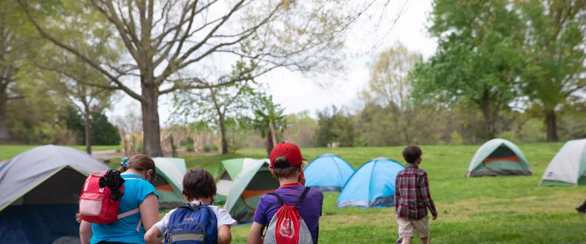 Four kids with backpacks hiking towards their pitched tents in a vast field that is sprinkled with trees at a NC Zoo camp.