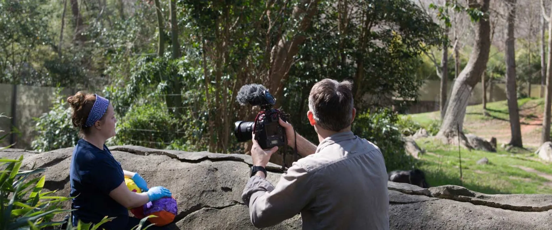 A female zookeeper stands next to a man who appears to be filming her interacting with an animal habitat. There is a large rock wall in front of them, overlooking a grassy field that is sprinkled with trees and boulders throughout the background.