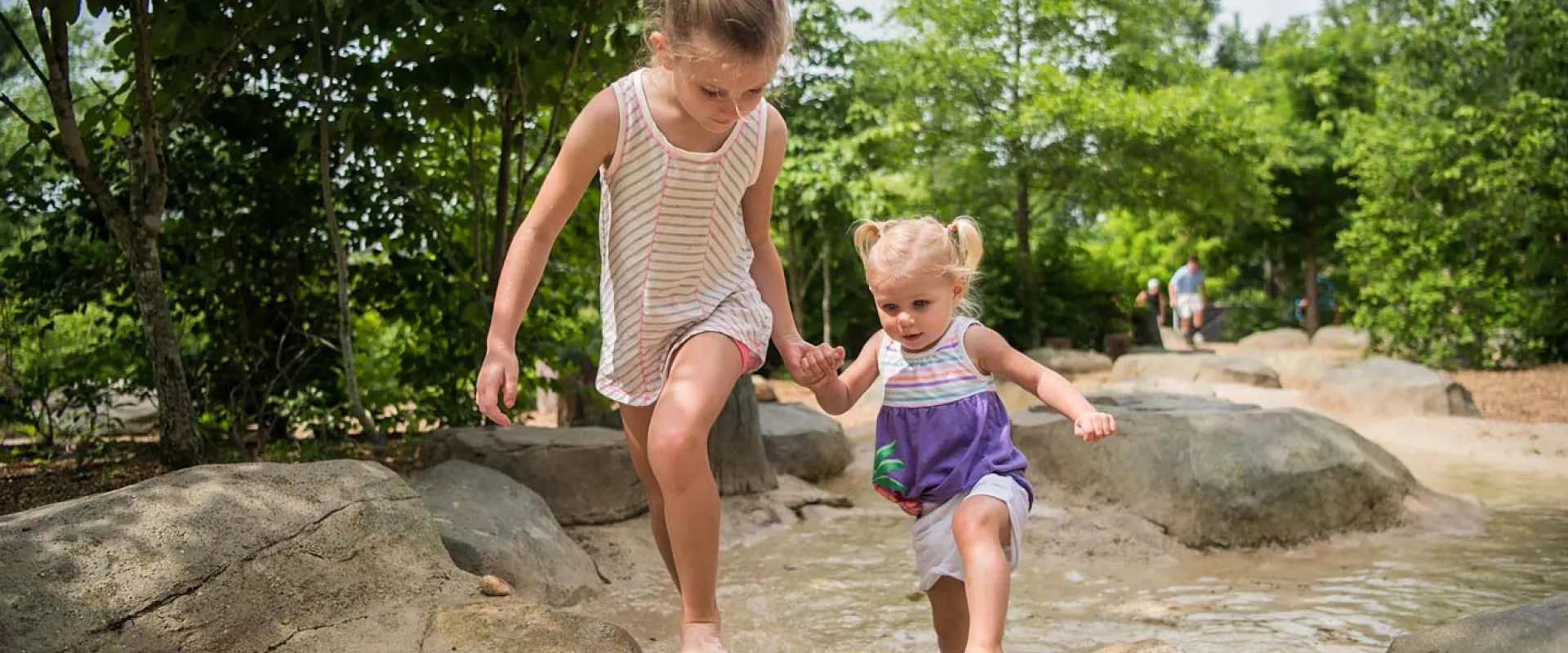 A young girl holding hands with a toddler as they climb barefoot up a steep, rocky hill. They are surrounded by trees and large rocks.