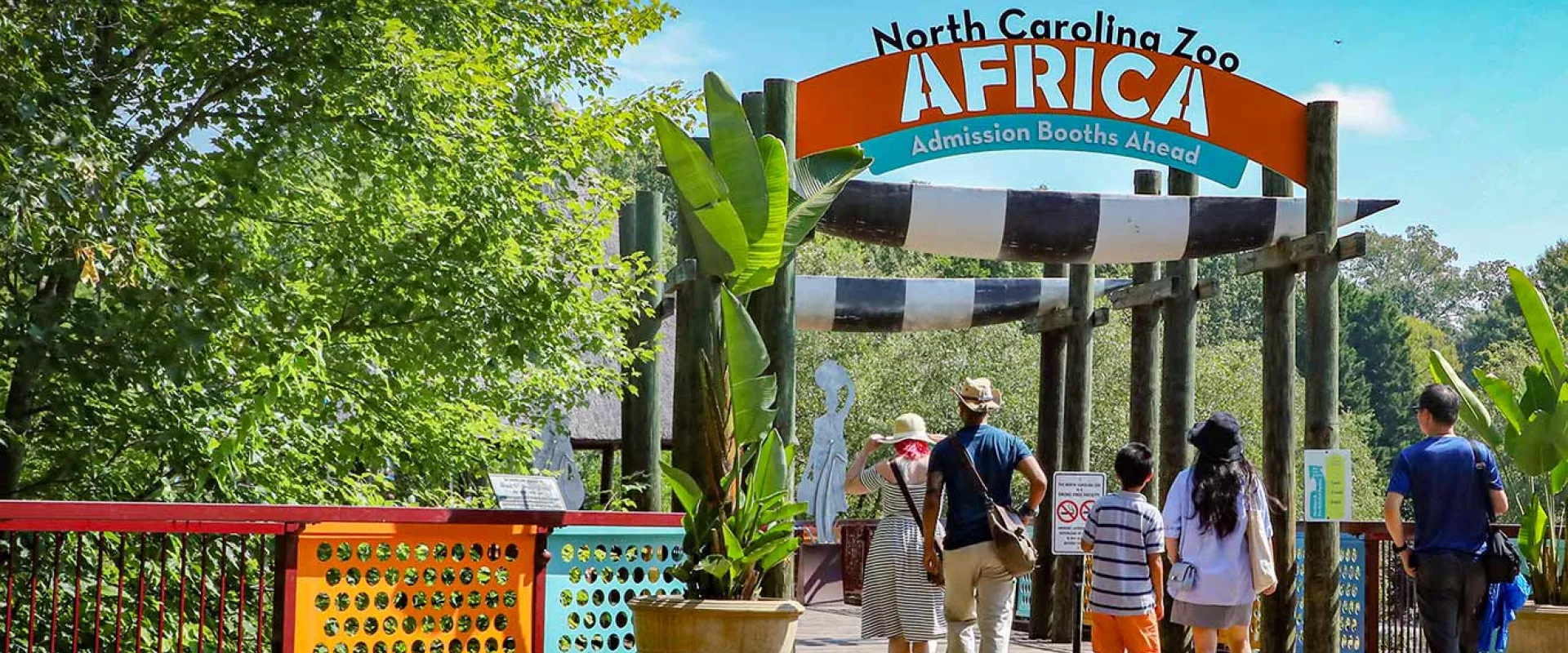 A wooden entrance sign, spanning a walkway, has a black and orange banner that reads "North Carolina Zoo AFRICA" with "Admission Booths Ahead" underneath. The walkway is bordered on the left by a brightly colored decorative fence with yellow, red, and blue panels. Several peopleare walking towards the entrance under the sign. The area is surrounded by lush green trees and tropical-looking plants in a terra cotta planter.