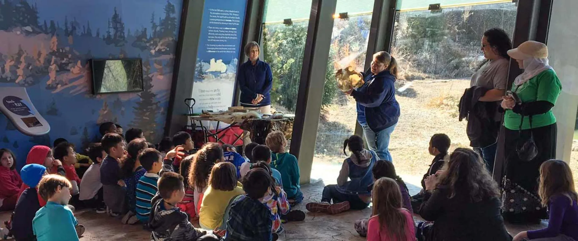 A group of approximately twenty children is sitting on the floor in a educational space with slanted glass windows. Two adult educators are leading the activity; one stands near a table with materials on the left, and another is kneeling to the right, showing the children an animal hide or fur. Large murals with landscape images and informational text are visible on the back walls.