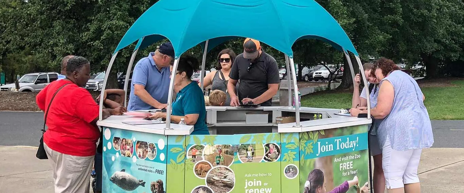 An outdoor zoo membership or information kiosk with a turquoise canopy. The kiosk is staffed by zoo employees and several guests are gathered around it. The sides of the kiosk feature images of animals and people with the phrase "Join Today!" and an invitation to renew membership.