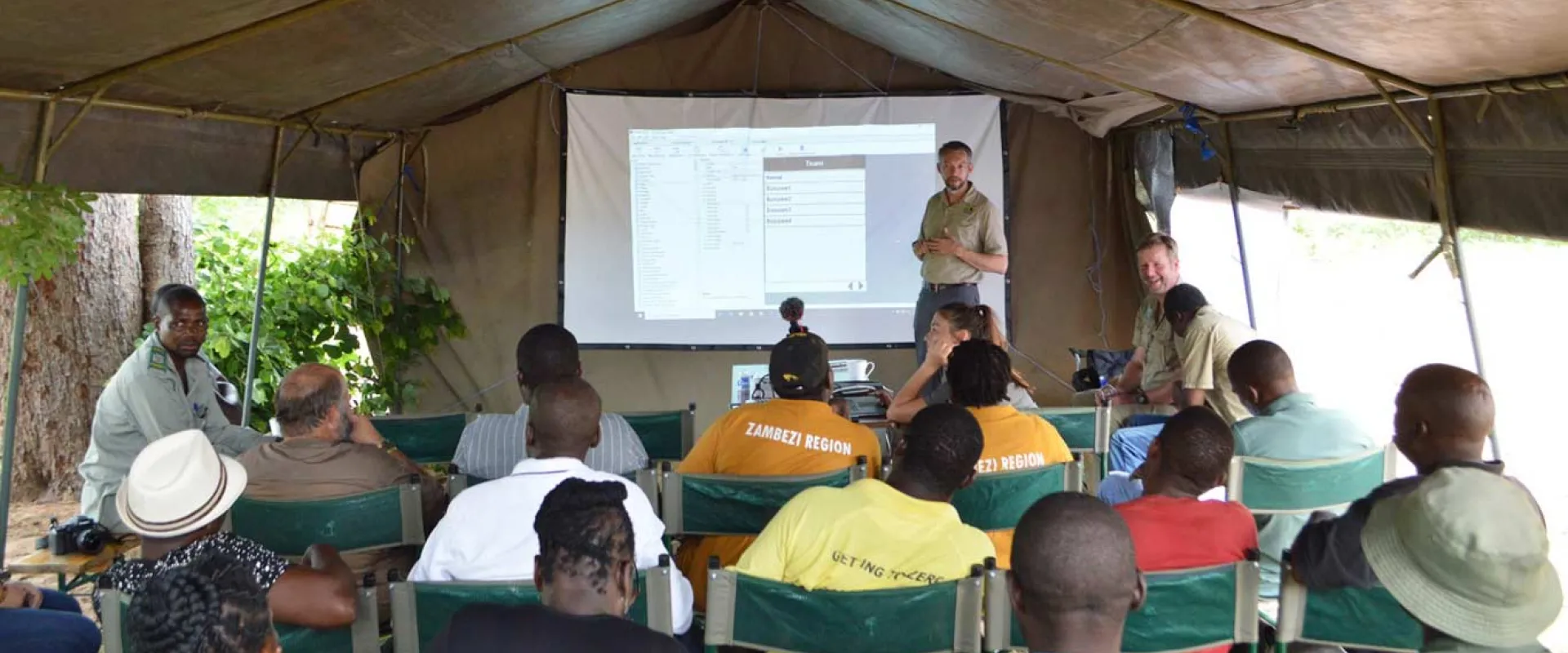 A large outdoor canvas tent, where a group of people are attending a presentation or training session. A man in a khaki shirt is standing under a projected screen on the back wall, speaking to the seated audience. About a dozen individuals, mostly men, are seated on metal-framed chairs, facing the front.