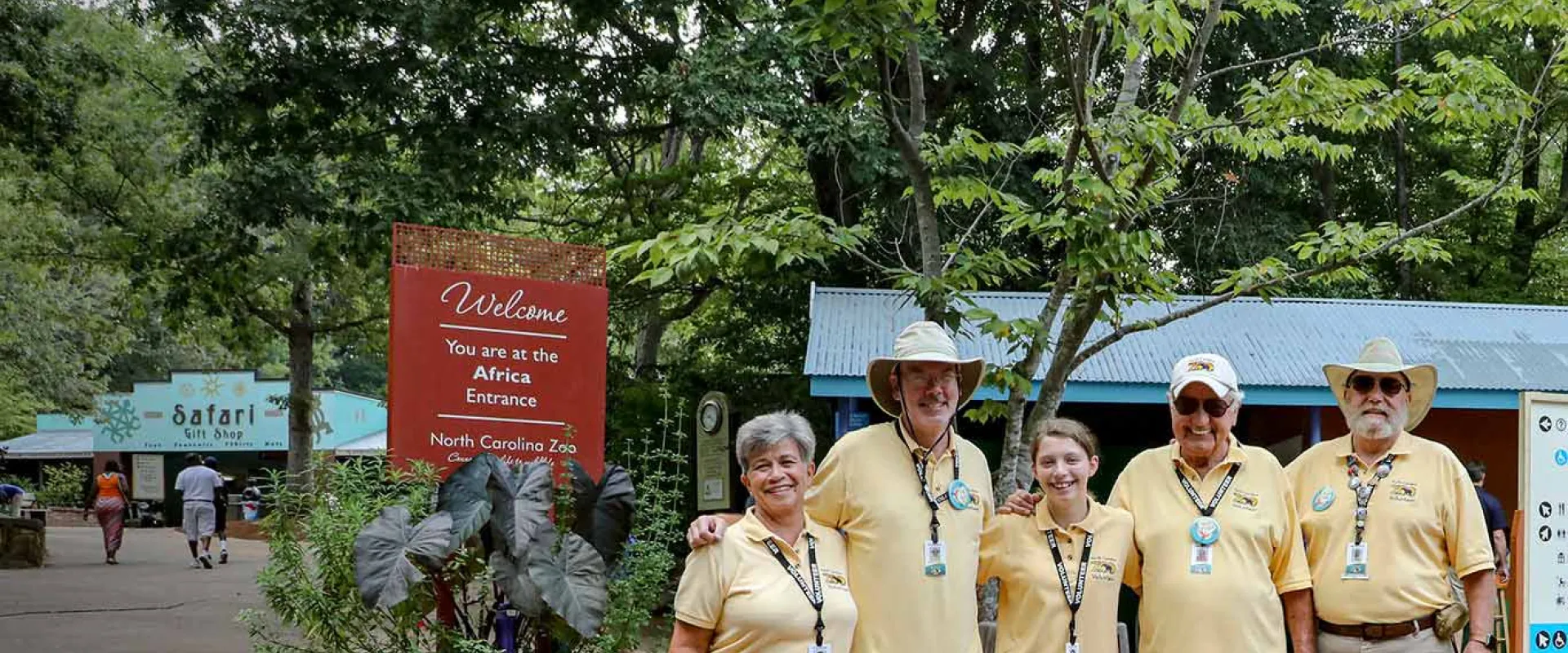 Five Zoo volunteers wearing yellow shirts and black lanyards posing, arms around each other, for the camera in the lush Africa Plaza.
