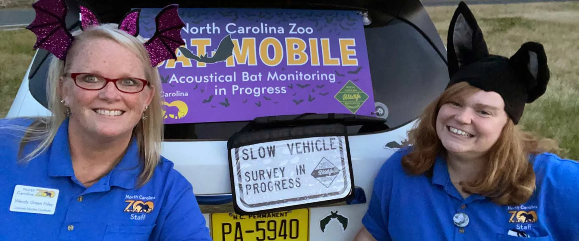 Two smiling women wearing blue shirts and novelty animal ear headbands (one with purple bat wings, one with black pointed ears) stand in front of the open trunk of a vehicle. A large purple and yellow sign reading "MOBILE" is visible in the trunk, likely for an outreach or education program.