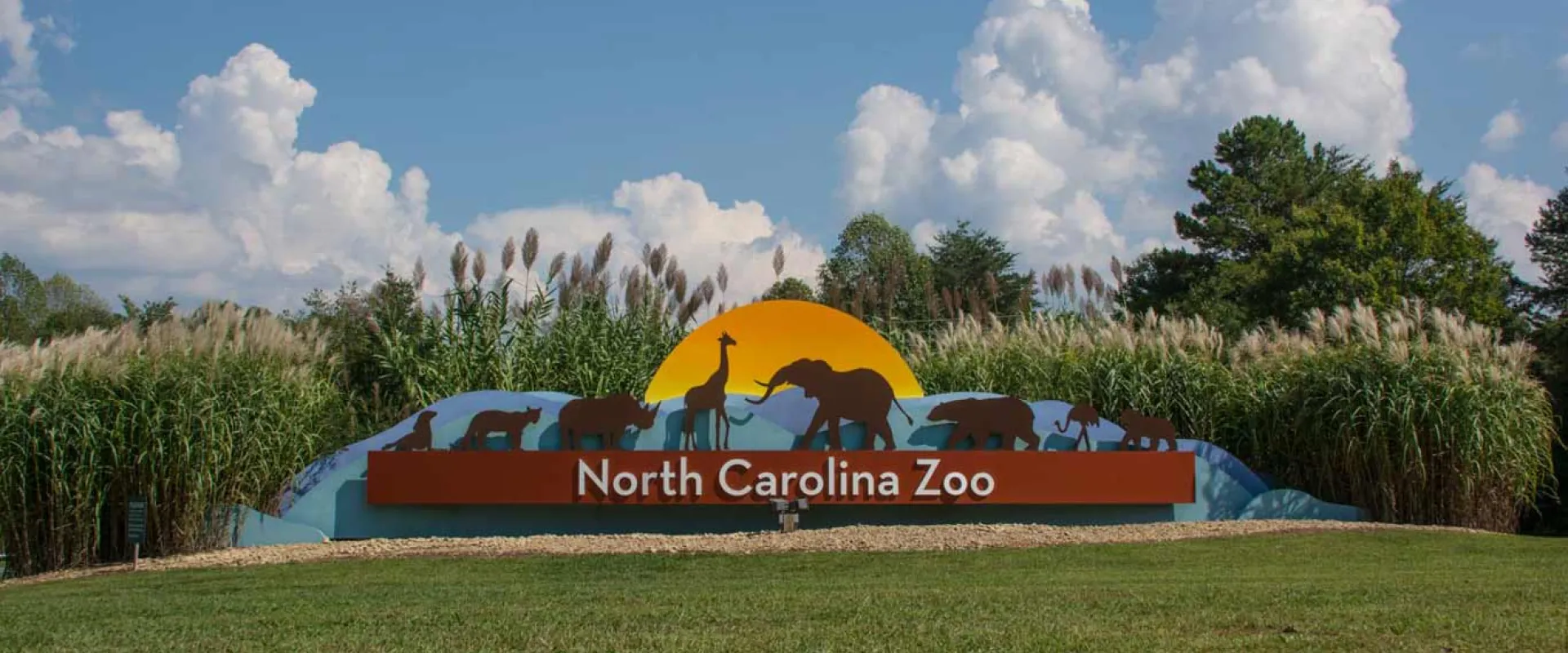 North Carolina Zoo Entrance Sign with animal silhouettes and colorful shapes and sun behind the animals.
