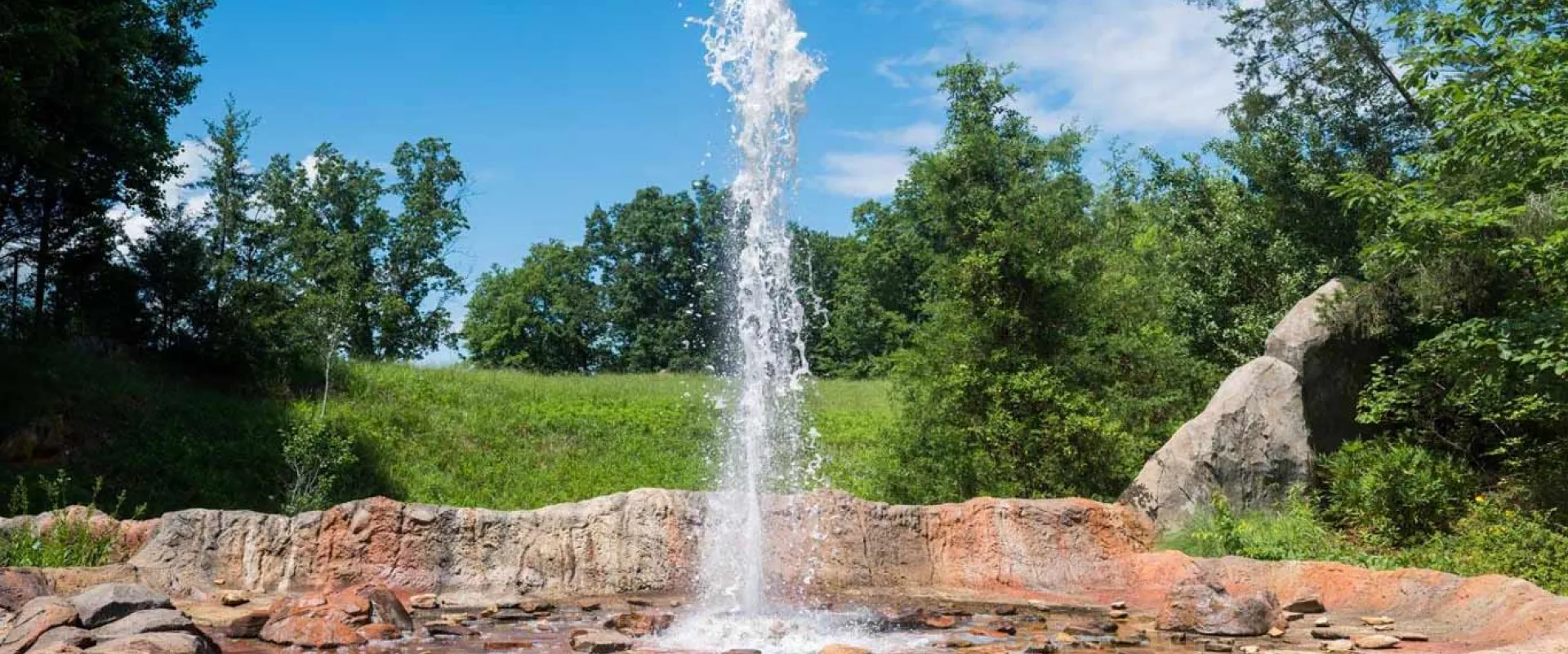 A fountain or geyser feature spraying a tall column of water high into the air against a bright blue sky with scattered white clouds. The water erupts from a base of reddish-brown and tan rocks, which are wet and surrounded by smaller, dark stones. The background is a vibrant green landscape with lush trees and a grassy hillside under the sunny sky.