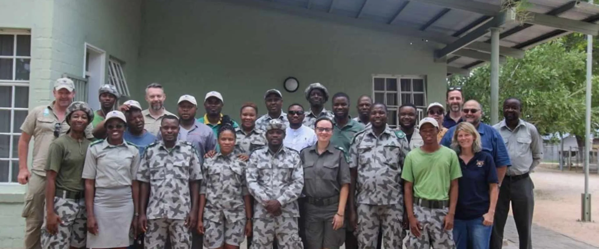 A large group of approximately 20 people, consisting of men and women, pose together for a group photo in front of a light green, single-story building. Most of the individuals are wearing matching camouflage or light-colored ranger/conservation uniforms, with a few wearing other casual outdoor attire. They appear to be a team of park rangers or conservation staff.