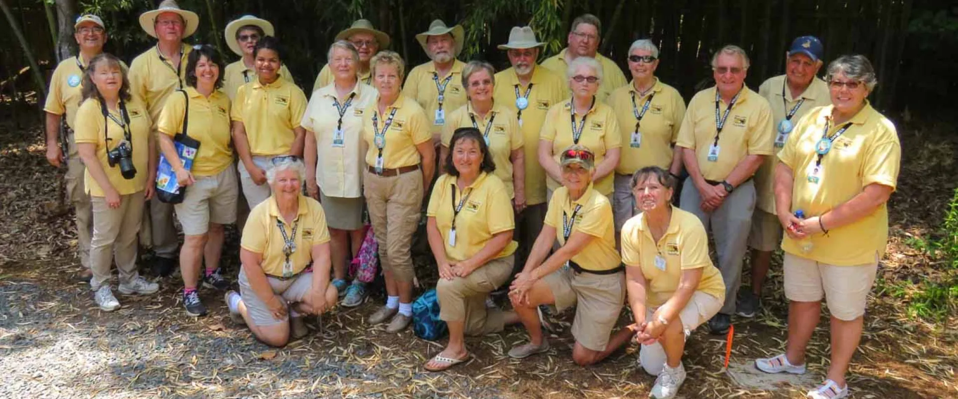 A group of approximately twenty adults, all wearing yellow polo shirts, stands together posing on a gravel parking lot with a dense forest in the background.